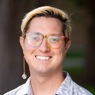 Zach Earl, They have colorful rimmed glasses, one long earring, short blonde hair, and a floral pattern shirt smiling at the camera